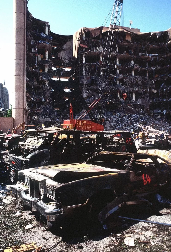 The bombed remains of automobiles with the bombed Federal Building in the background.