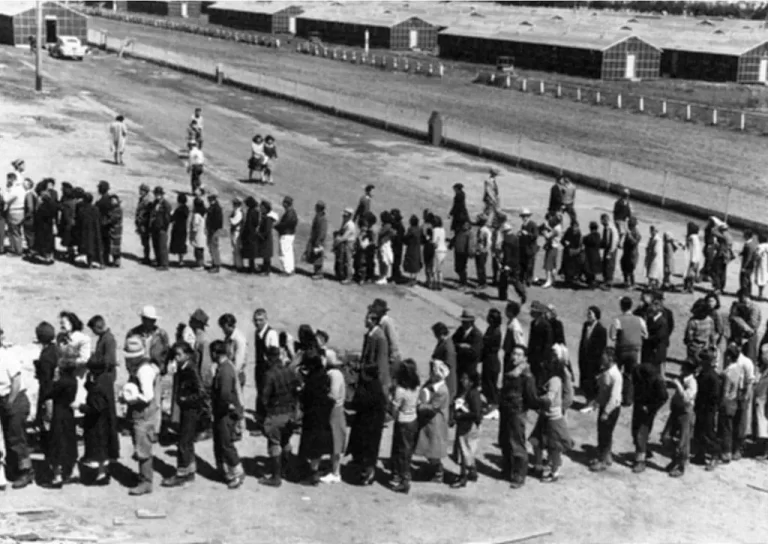 A Japanese-American internment center. Description: This assembly center has been open for two days. Only one mess hall was operating today. Photograph shows line-up of newly arrived evacuees outside of this mess hall at noon. Tanforan Assembly Center. San Bruno, CA, April 29, 1942. Dorothea Lange.