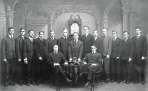 15 Bonesmen and their grandfather clock, group portrait, black and white. The Skull and Bones has a private group portrait taken for every new cohort of fifteen. It is always posed in the same manner, showing human bones and a grandfather clock at 8 p.m.